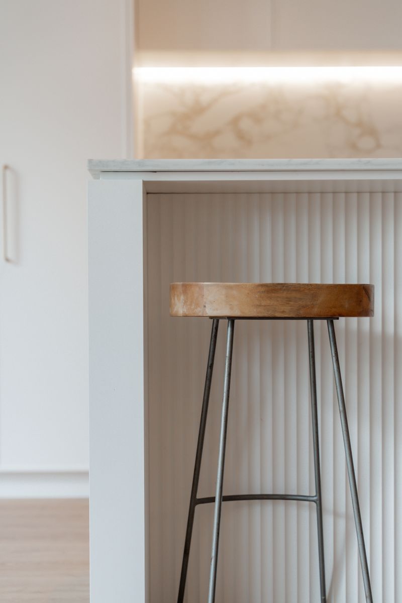 Timber bar stool against the white fluted island, with the lit splashback behind