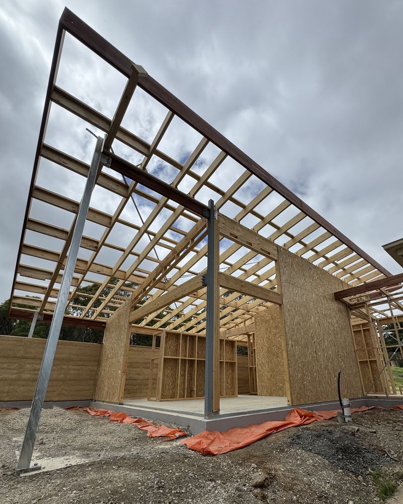 Looking up from a corner of the Willunga addition mid-build: galvanised steel posts and beams carrying open timber roof framing, braced ply walls and the rammed-earth walls beyond