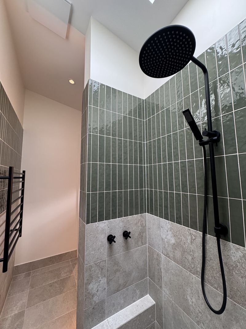 Walk-in shower with glazed green vertical tiles above stone-look porcelain, a matte black rain shower and a skylight above