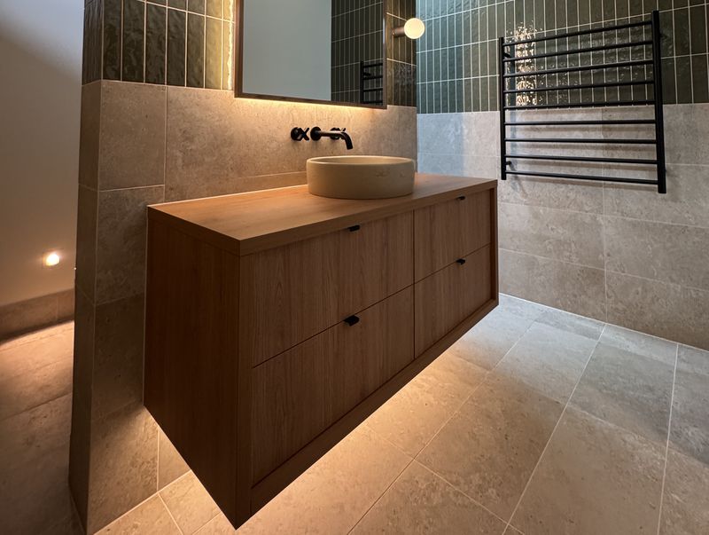 Floating timber vanity with a stone vessel basin and matte black tapware, softly lit from beneath, beside a heated towel rail